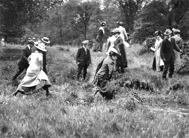 Black and white photograph of around a dozen people wearing hats, dresses, and suites as they cross a prairie stream