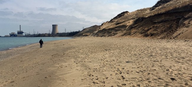 The shore in front of Mt. Baldy with NIPSCO's cooling tower in the background and man walking along the beach.