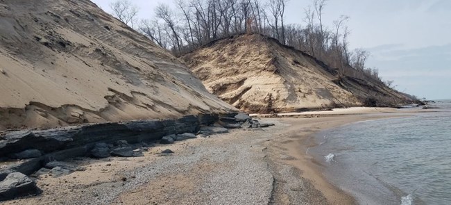 Outflow of a drainage ditch into Lake Michigan with dunes on either side of the stream and a layer of dark earth material exposed underneath the dunes.