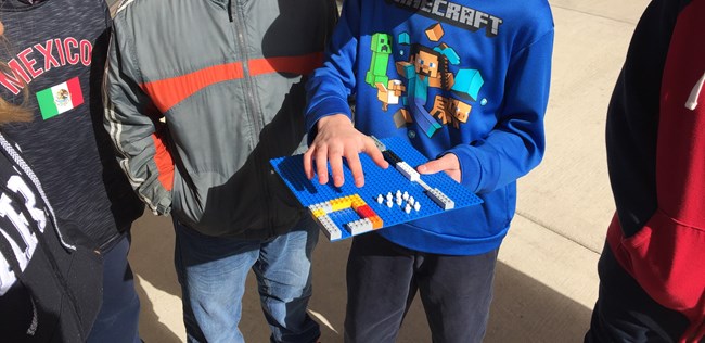 A student proudly holds their group's carp barrier design made from plastic toy construction bricks while the other four group members circle around.
