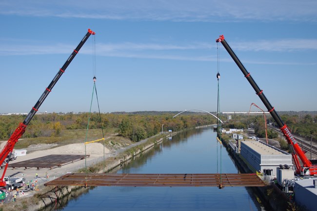 Two large cranes straddle the opposite edges of a waterway. Suspended by the cranes above the water is a flat, rectangular, rusty metal grate structure that is being lowered into the water.