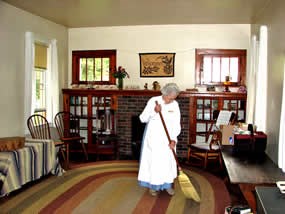 room with rag rug on floor with lady sweeping it, windows and bookcases on wall on both sides of a fireplace.