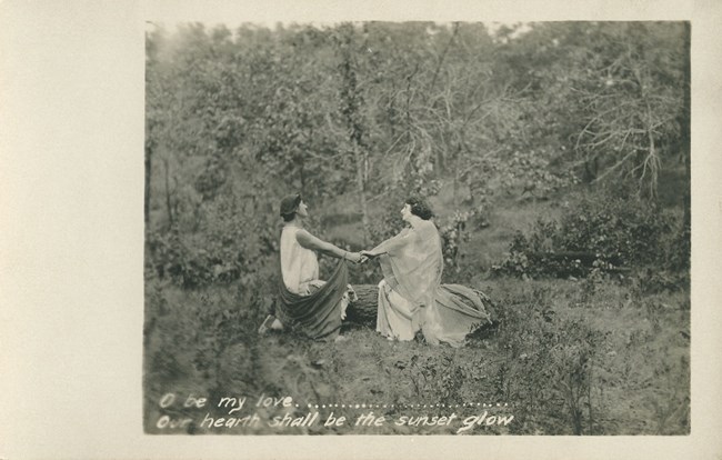 Historic sepia photograph from 1913 of a masque performance in the Indiana Dunes. Performers, man and woman, hold each other's outstretched hands among a forest scene.