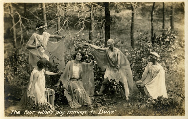 Sepia photograph from 1913 of a scene from historic masque entitled "Duna the Spirit of the Dunes." Characters in costume with outstretched arms surround a woman seated in nature.