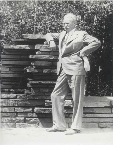 Jens Jensen, a tall man in a suit with white hair and a white mustache stands leaning on fountain built of stacked stone tablets. Black and white.