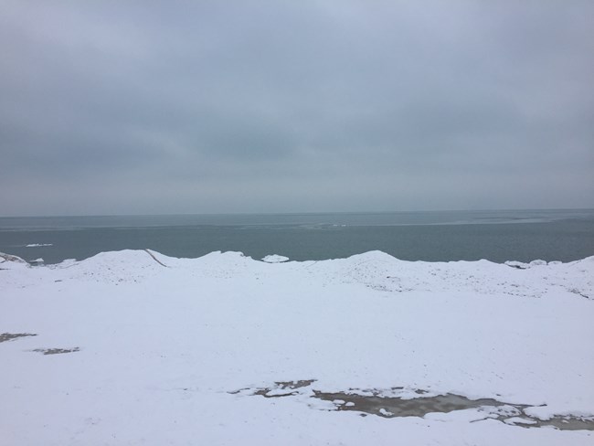White snow and ice line the foreground, beyond the ice, Lake Michigan's open water appears gray blue below and gray sky.