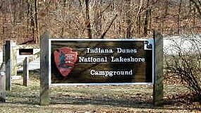 a carved large wooden sign with NPS arrowhead and woods Indiana Dunes National Lakeshore   Campground raised above plain background in front of spring trees