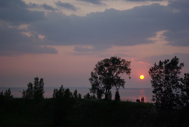 People on the beach watching the sun set over the lake between two trees. The sky is pink, blue, and purple.