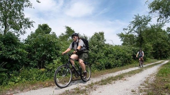 Two rangers ride bicycles down a gravel trail alongside shrubs and trees.