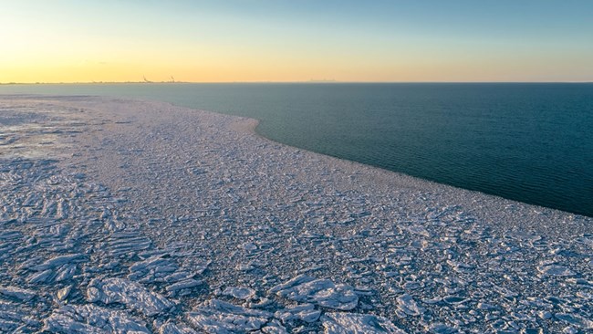 Aerial view of Lake Michigan in winter. Open blue water on right. Fragmented, inconsistent hunks of ice fuze together along the shore.