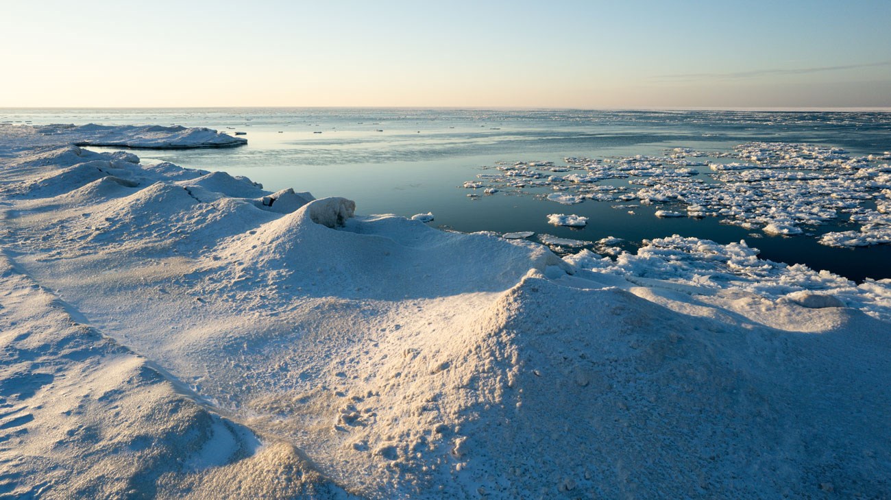 White hills of ice line the shore of Lake Michigan. Blue waters with floating ice in background. Sky is blue with yellow on the horizon.