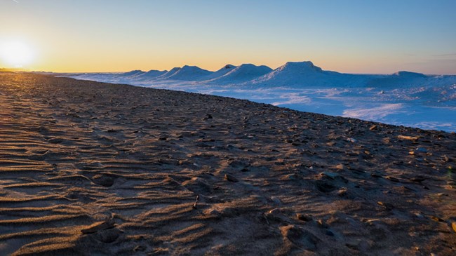 White mounds of ice rise against a sandy beach. A soft yellow sun sets on the left.