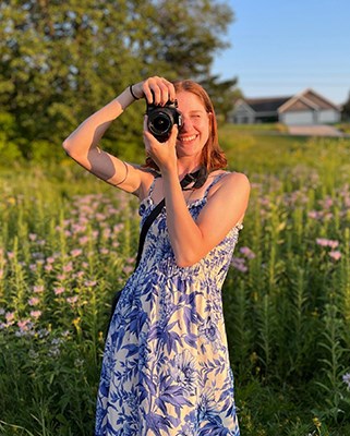 portrait of artist holding a camera to her eye in front of green field with house in far background