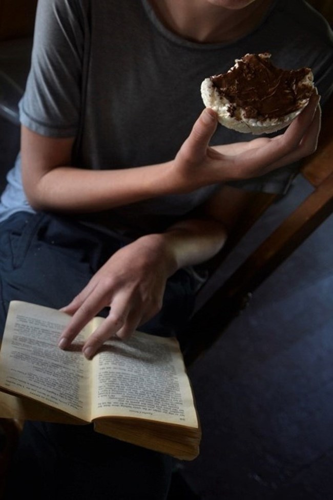 person holds half-eaten rice cake with some type of dark topping, perhaps hazelnut spread, while holding book open with other hand