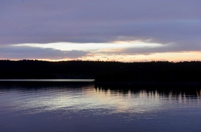 darkness stretches across the treed shoreline, waters of the lake dark, the sky cloudy with oranges and yellows