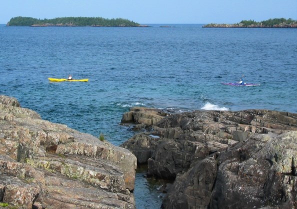 A yellow and purple kayak are on the water with islands in the background. Photo taken from shoreline in foreground.