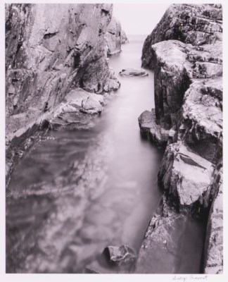 A black and white image shows a narrow break in cliffs with water in the middle