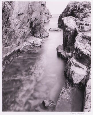 A black and white image shows a narrow break in cliffs with water in the middle