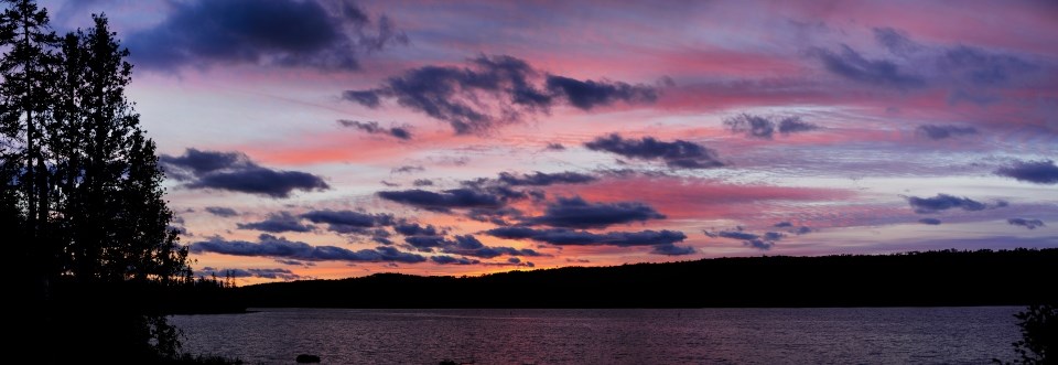 A panoramic image shows a pink, blue, and purple sunset over a large body of water lined with trees