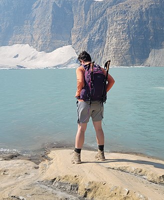 artist standing on a rock facing a light blue lake with snow covered mountains in the distance
