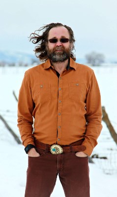 A man with flowing hair and sunglasses stands on snow. He has red pants and an orange long sleeve shirt on
