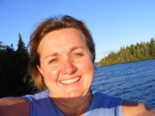 A woman smiles at the camera at close range with water and trees in the background against a sunny blue sky