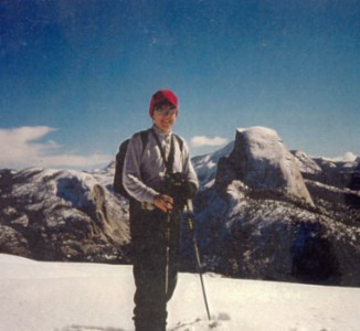 A woman stands on a snowy peak with large mountains and blue sky in the background. She has ski poles in her hands, a winter hat, and a backpack on.