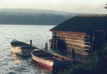 A historic wood boathouse is on the right and sits right on the lake, two small boats sit on a dock attached to the boathouse. A near shore is visible across from the boathouse.