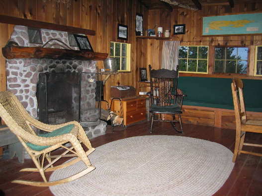 A rustic cabin with a large stone fireplace, chairs, and a couch surrounding a large circular rug