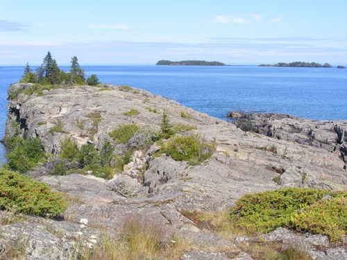 A photograph shows the end of a rocky peninsula surrounded by a bold blue lake with small islands in the far distance