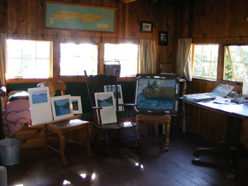 A photograph shows the inside of a wood cabin with six small paintings sitting on chairs leaning against the backs of them. Sunlight blasts through the windows