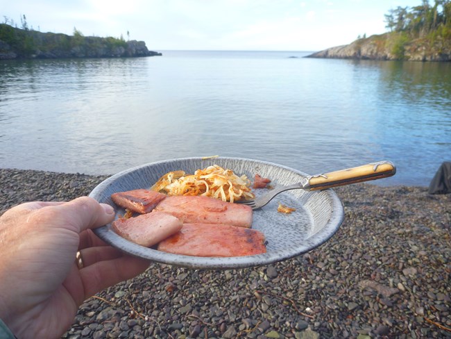 A person holds up a breakfast plate consisting of ham and hashbrowns while camping with a lake and stone beach in the background.