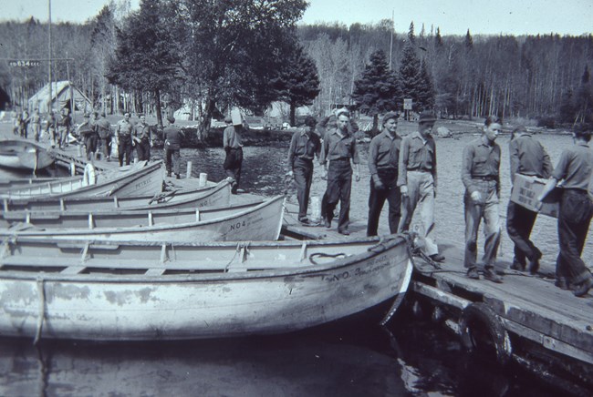 An old, black and white photo, shows lines and men walking up and down a dock lined with small white boats, carrying supplies to their camp.