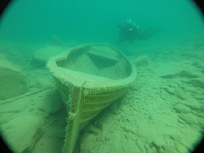 A small boat at the bottom of a lake with a scuba diver nearby.