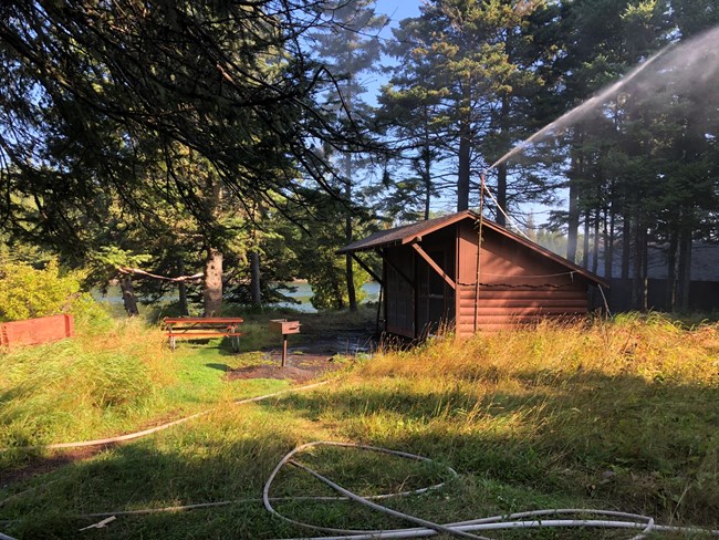 lt= A tall water sprinkler set up next to two brown three sided camping shelters, spraying water around the campground.