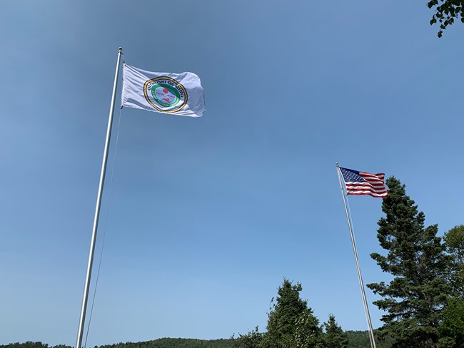 A United States flag and Grand Portage Band of Lake Superior Chippewa flag fly next to each other against a clear blue sky.