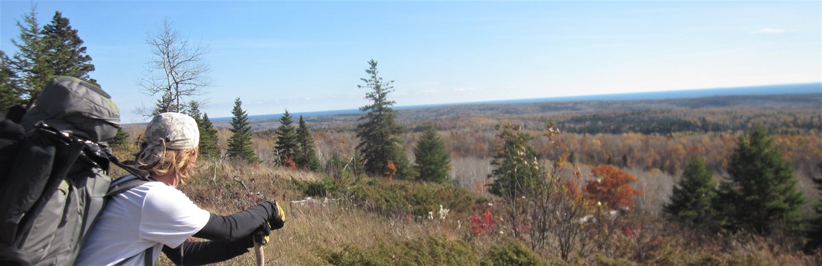 A person with a backpack stands on an exposed rocky ridgetop overlooking shrubs, forest, and a lake.