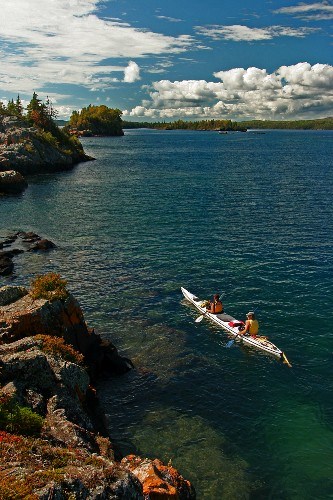 Two people in a long white sea kayak paddle on Lake Superior near Isle Royale's rocky shore.
