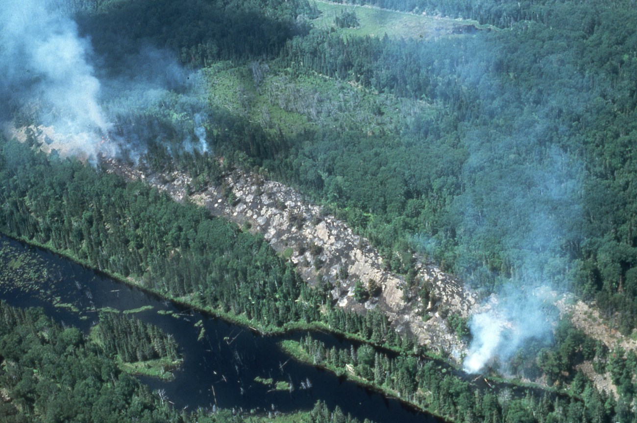 A grainy, vintage aerial photo shows two separate plumes of smoke rise from a forest.