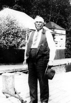 captain standing in front of lodge on dock while smoking a pipe and holding a tophat in left hand