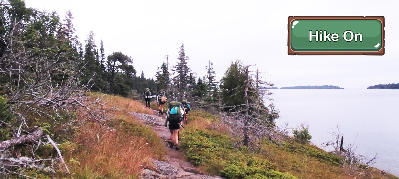 People carrying large backpacks hike on a lakeside, rocky trail.