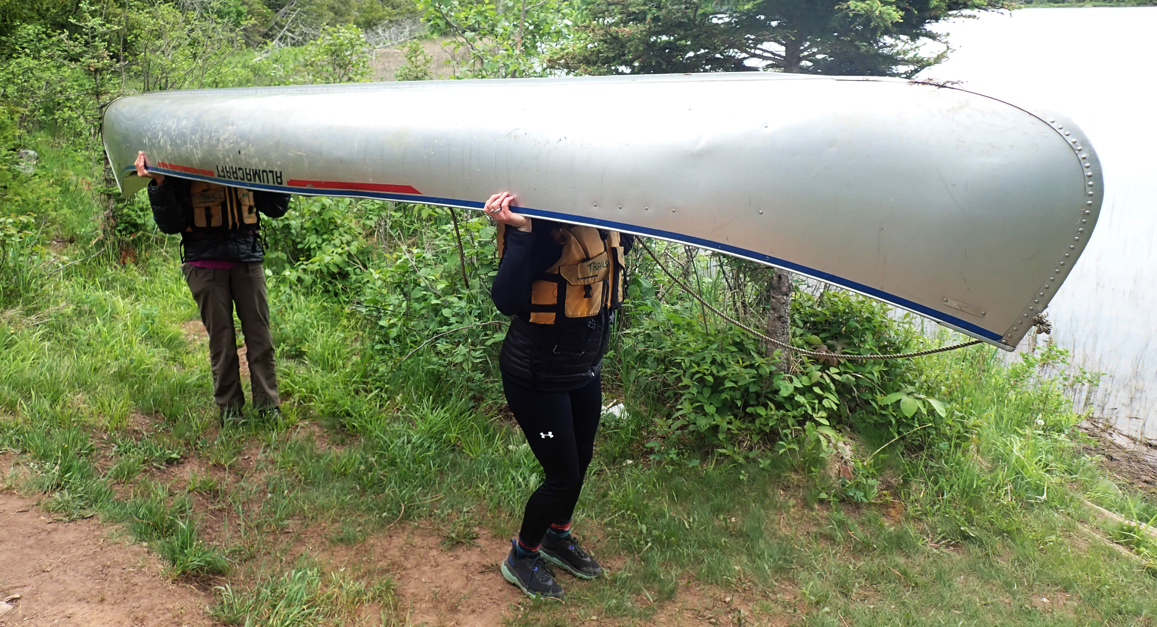 Two people carry a canoe by a lake.