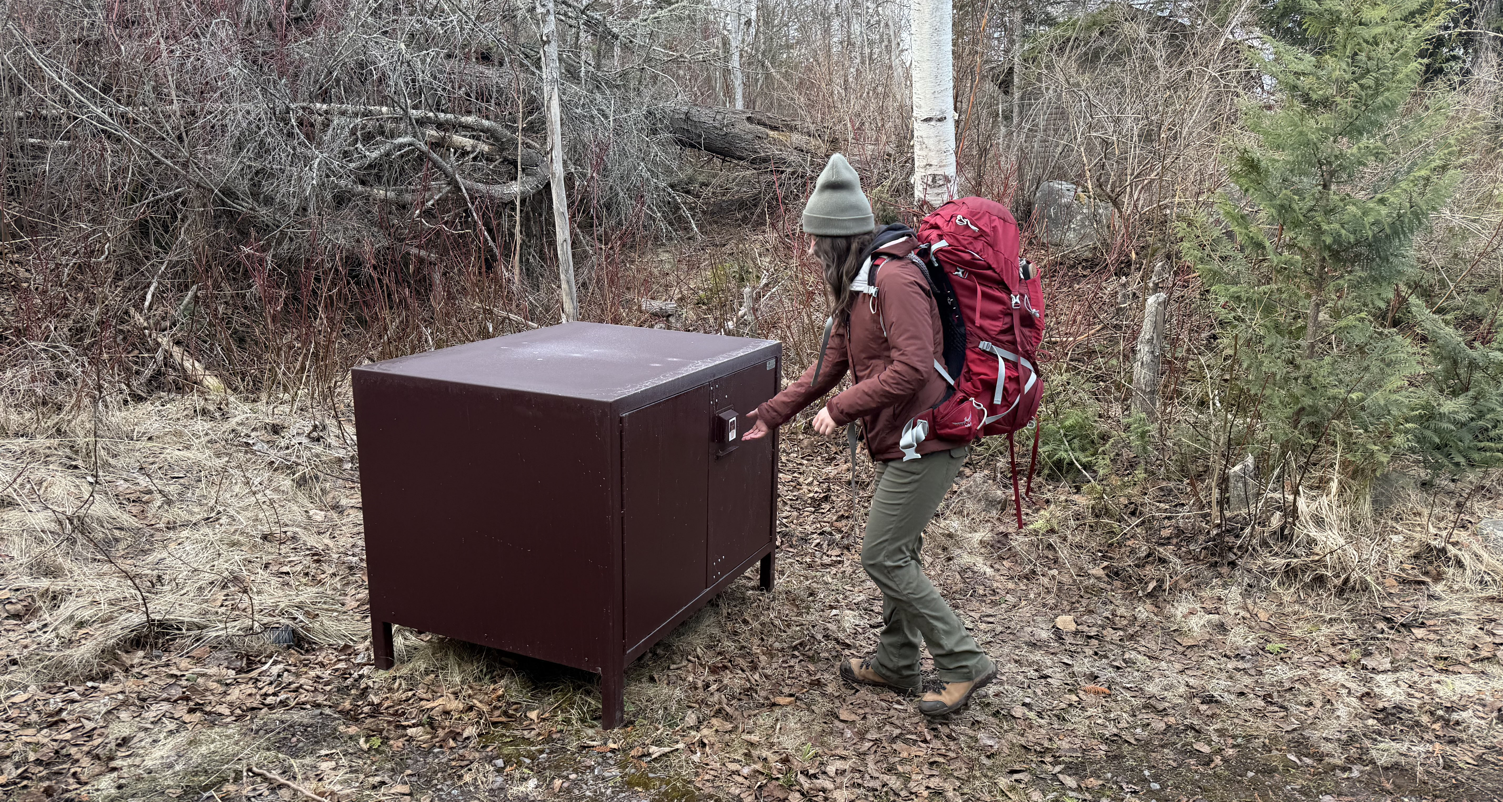 A person wearing a large red backpack, a red coat, green pants, and a green hat approaches a large, brown, metal food storage locker in the woods.