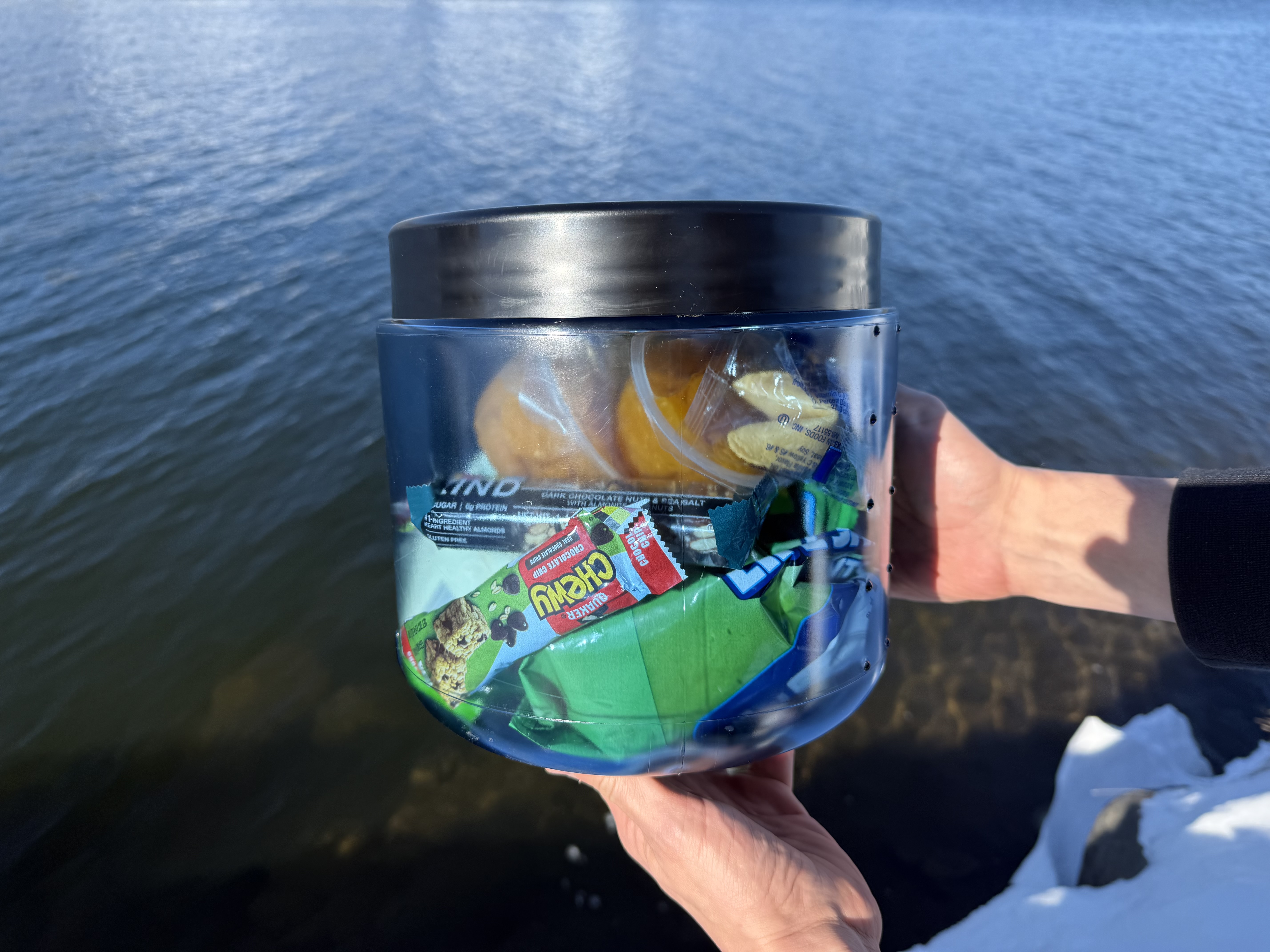 A round, blue, see through container with a black lid, containing multiple snacks and food items.