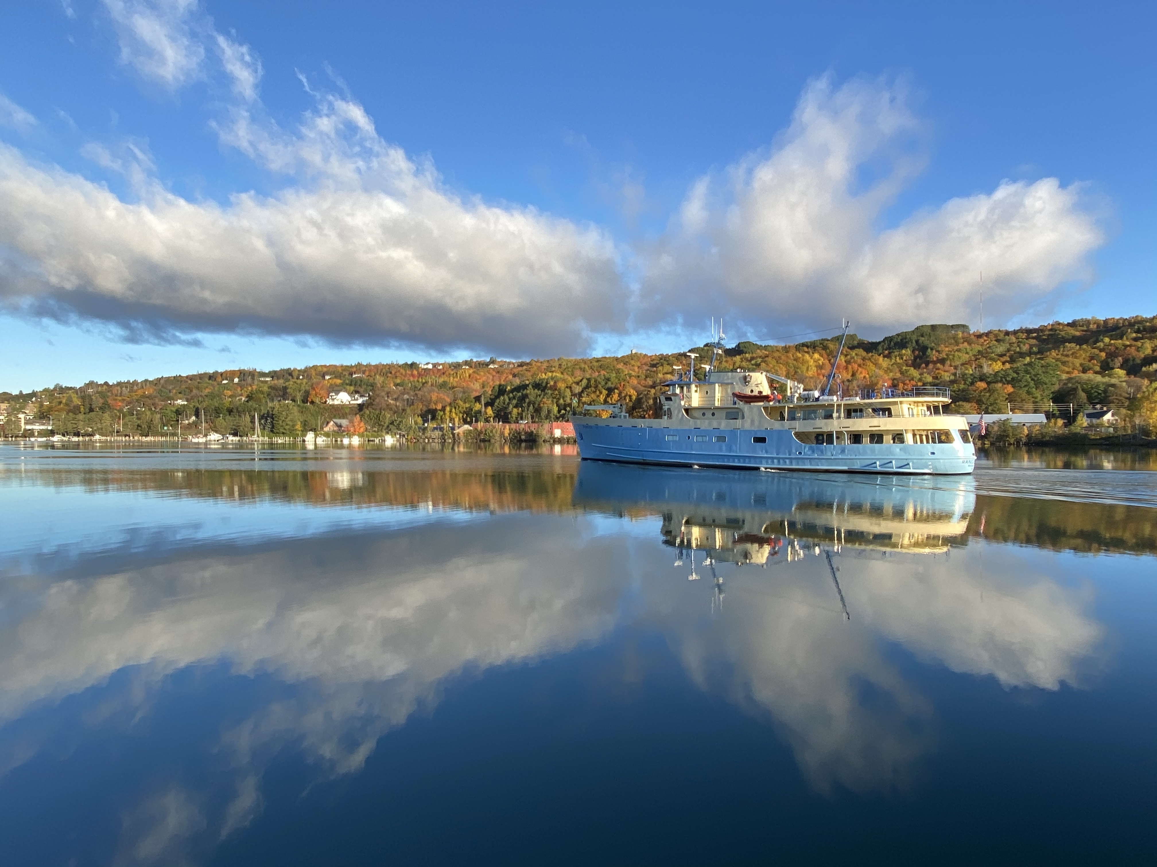 A large, blue and yellow ferry sails in a tree lined canal.