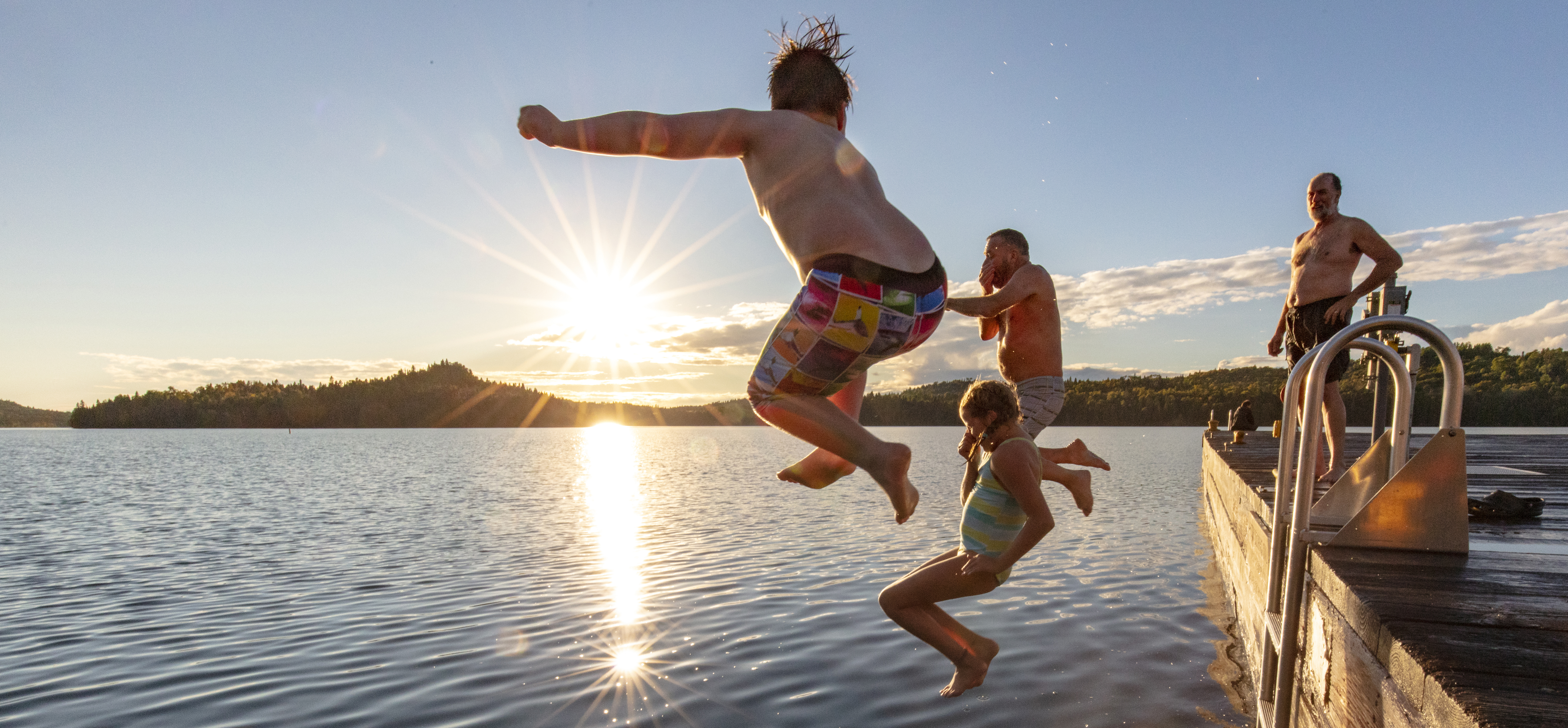 Three people jump off a dock and into a lake while a fourth person stands on the dock and watches.