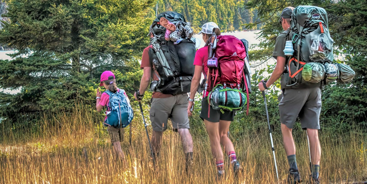 Four people with backpacks hike in all grass with a forest and water in the background.