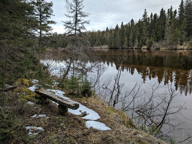 Snow beneath a bench next to a river.