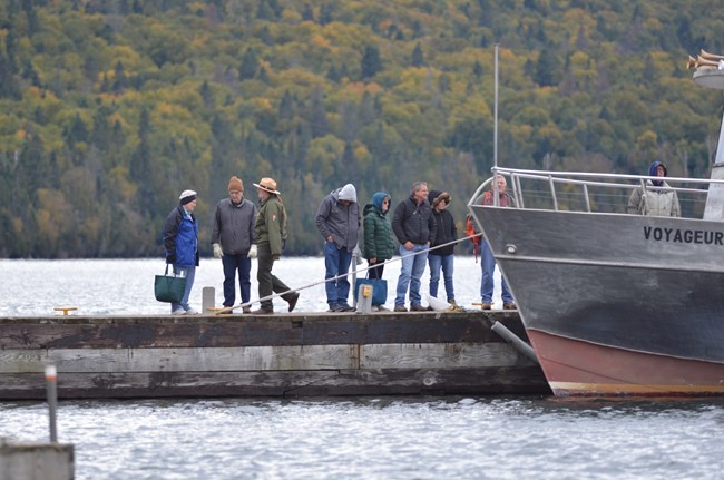 A ranger walks down a dock lined with people in wiinter coats waiting to get on a ferry. The trees in the background have turned orange.
