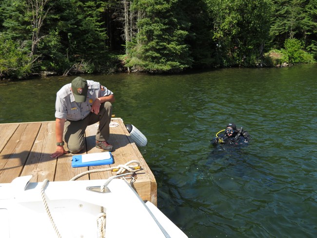 A person kneels on a dock taking notes on a clipboard, while a diver is in the water beside them.